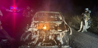 A soldier shines a flashlight at an incinerated car at night