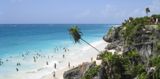 Tourists follow a staircase down a cliff to a white sand beach with turquoise water