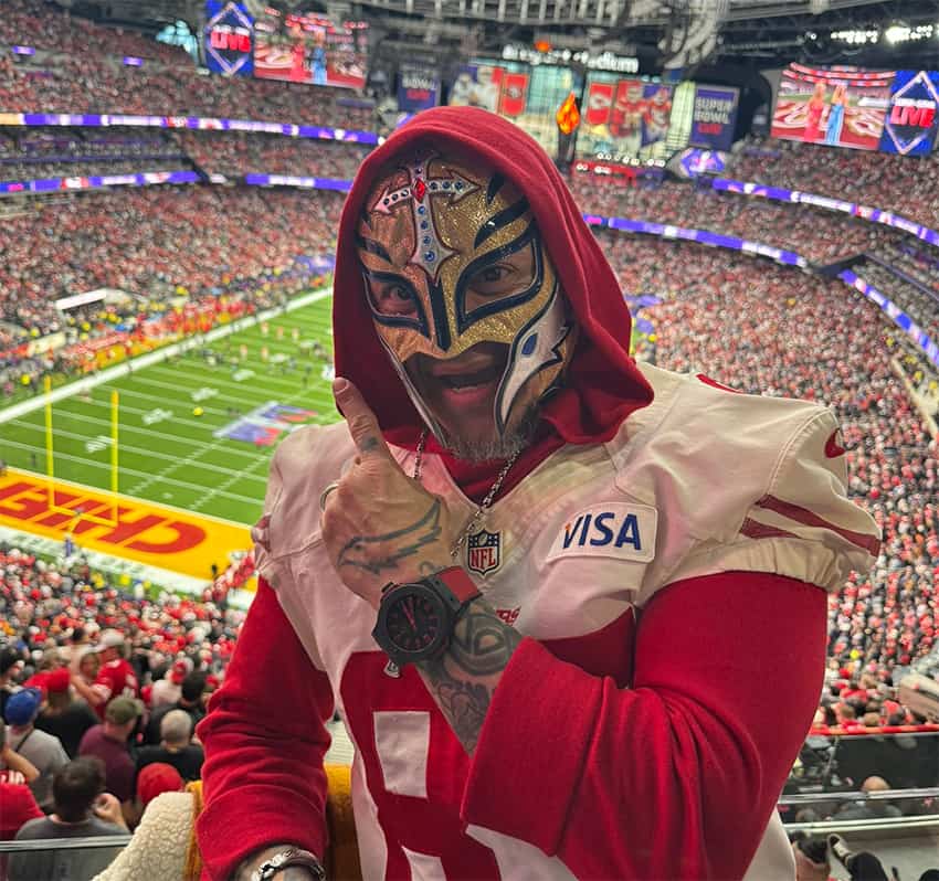 A pro wrestler in a lucha libre mask poses with a full stadium and football field behind him