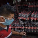 A man wearing a facemask unloads packages of plastic Coca Cola bottles from a truck