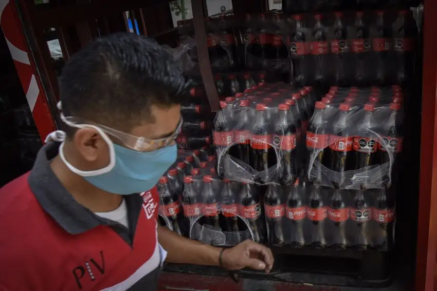 A man wearing a facemask unloads packages of plastic Coca Cola bottles from a truck