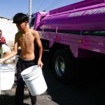 A young man in Mexico City neighborhood holding several large buckets in front of a city potable water distribution truck