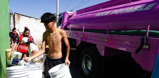 A young man in Mexico City neighborhood holding several large buckets in front of a city potable water distribution truck