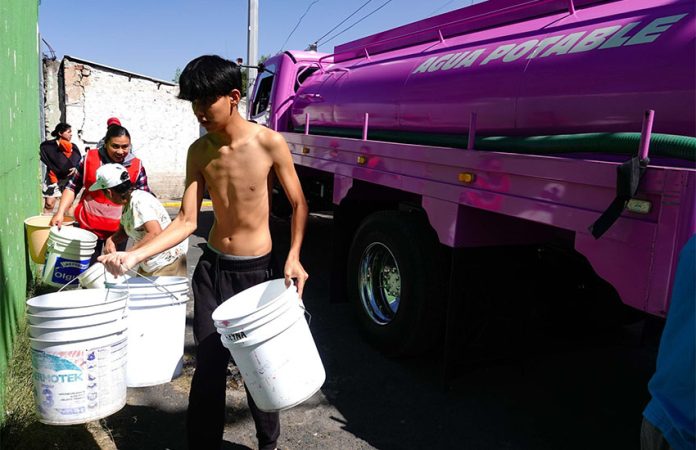A young man in Mexico City neighborhood holding several large buckets in front of a city potable water distribution truck