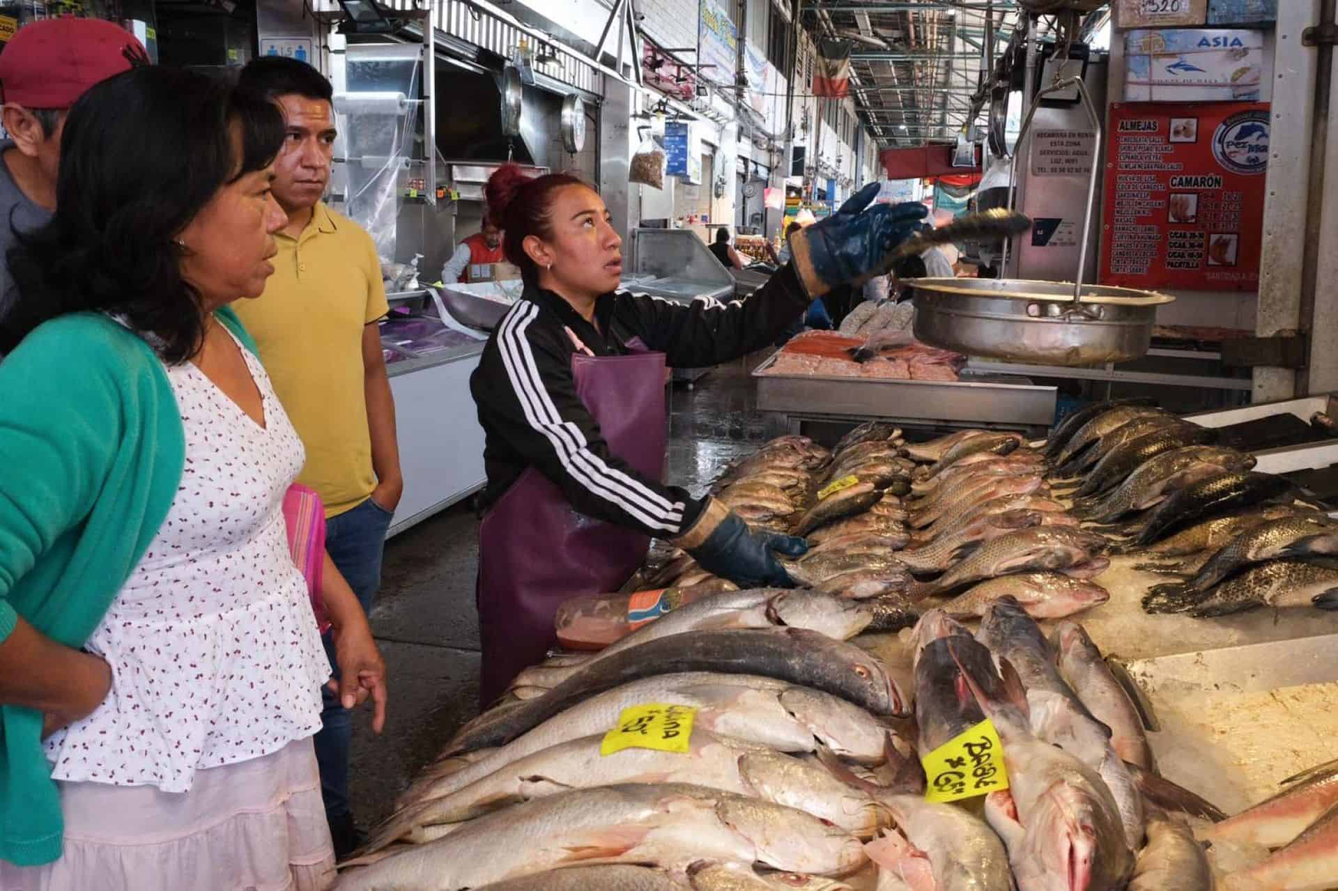 A female fishmonger weighs fish on a hanging scale for a male and female customer
