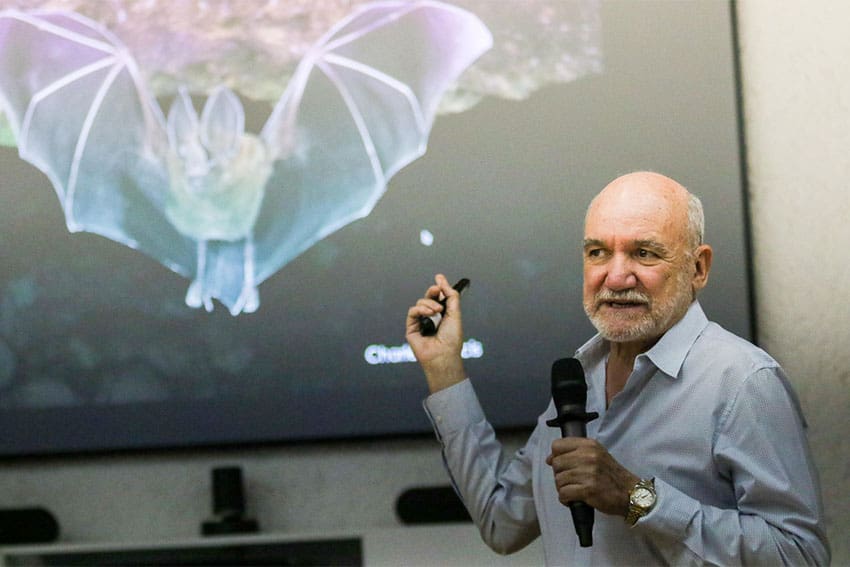 Bat expert Rodrigo Medellin with mic in hand, in front of digital display of a bat