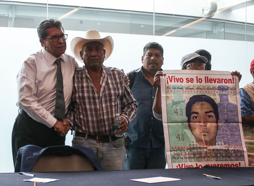 Ayotzinapa students' fathers posing with Mexico's Senate Majority Leader Ricardo Monreal, one holding a poster of his son