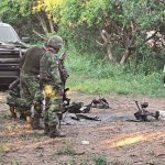 Soldiers look at spent explosives on a rural road, with their truck in the background.