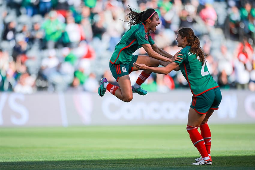 A soccer player in a green jersey jumps for joy and grabs her teammates hand, on a professional soccer field
