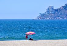 Acapulco beachgoer in front of ocean with damaged buildings in coastline view