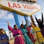 Six women in traditional Raramuri dress with raised hands under the Welcome to Las Vegas sign,