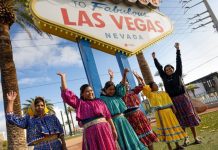 Six women in traditional Raramuri dress with raised hands under the Welcome to Las Vegas sign,