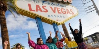 Six women in traditional Raramuri dress with raised hands under the Welcome to Las Vegas sign,