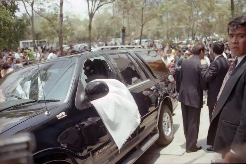 A large black SUV with its driver's side window shot out and men in suits standing around it