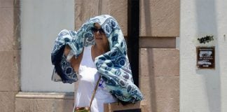 A middle-aged Mexican woman standing next to a building with a large blue and white scarf hanging over her head and shoulders to protect herself from high temperatures in Oaxaca city during a heat wave