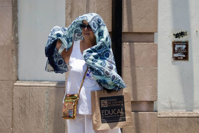 A middle-aged Mexican woman standing next to a building with a large blue and white scarf hanging over her head and shoulders to protect herself from high temperatures in Oaxaca city during a heat wave