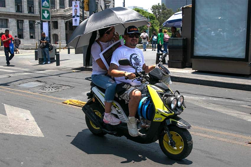 Two Mexican men on a moped driving through Mexico City streets and carrying an umbrella over their heads to shield themselves from the sun