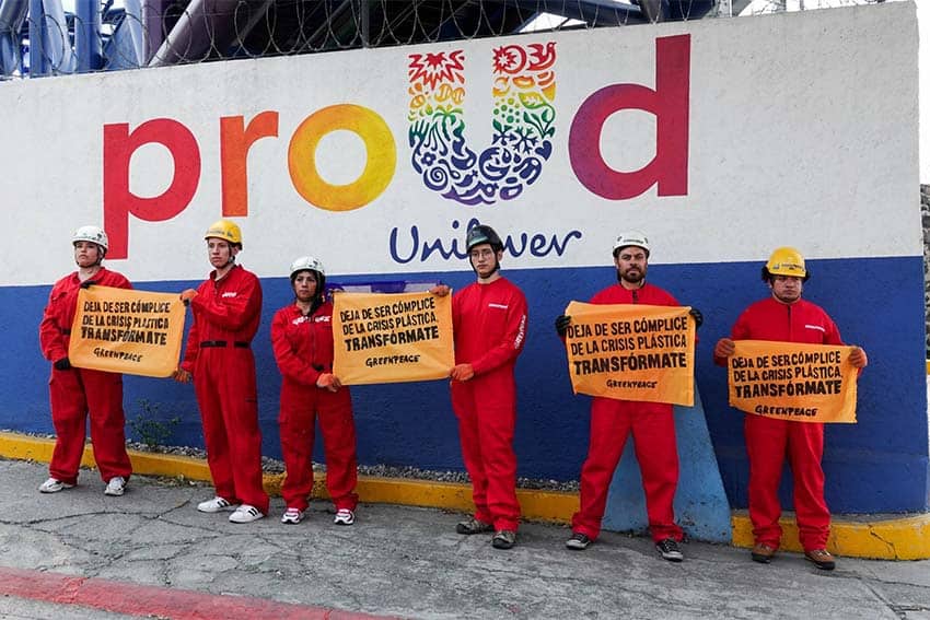 Six Mexican Greenpeace activists in hardhats and red work jumpsuiits with protest signs against plastics standing in front of a Unilever factory in Mexico