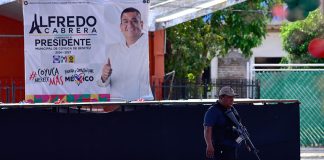 A federal agent patrols a plaza with a banner for murdered mayoral candidate José Alfredo Cabrera Barrientos in the background.