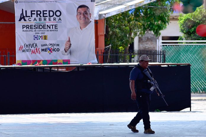 A federal agent patrols a plaza with a banner for murdered mayoral candidate José Alfredo Cabrera Barrientos in the background.