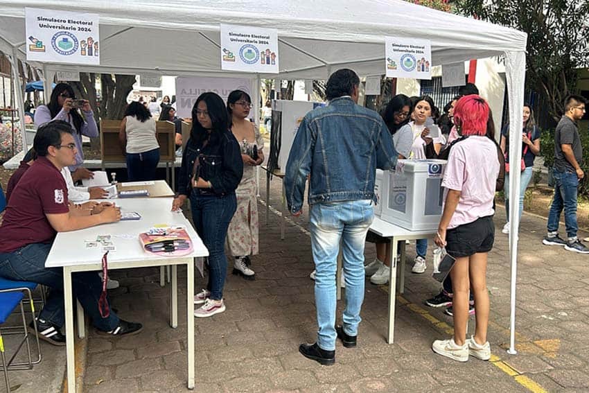 Colleges students voting in Mexico's University Electoral Simulation under outdoor tents with tables and ballot boxes