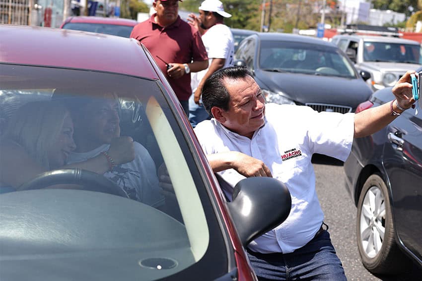 Yucatan, Mexico, gubernatorial candidate Joaquin "Huacho" Diaz standing outside a car in traffic taking a selfie with two supporters who are inside a car.