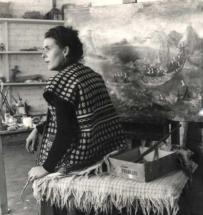 Black and white photo of artist Leonora Carrington with paintbrush in her hand, sitting on a table in a traditional huipil, near her painting on an easel