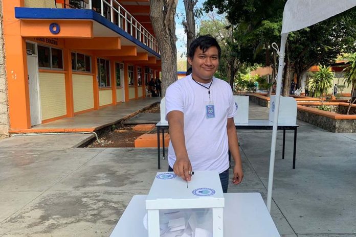 Smiling Mexican college student in a tee shirt and jeans placing a ballot in a box for a mock presidential election