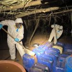 Two people in hazmat suits and with shovels removing industrial sized jugs of chemicals from a synthetic drug lab in Sinaloa