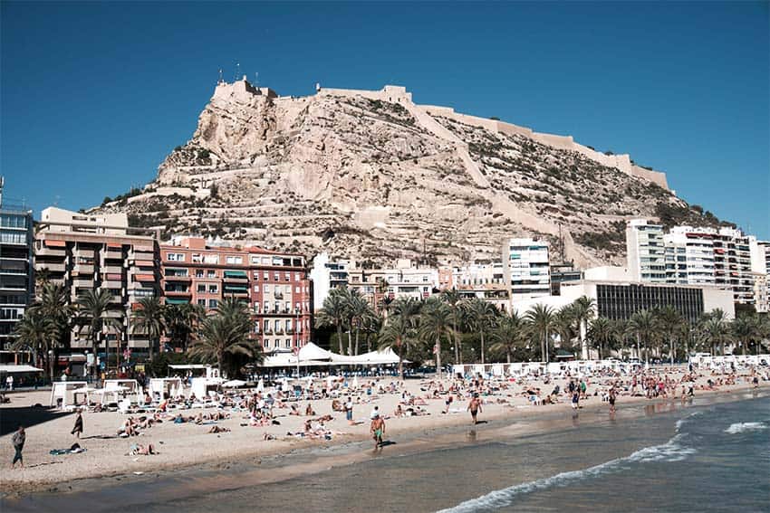 Crowds on the shore of a beach in Alicante, Spain, with buildings behind them and a large rock face in the background.
