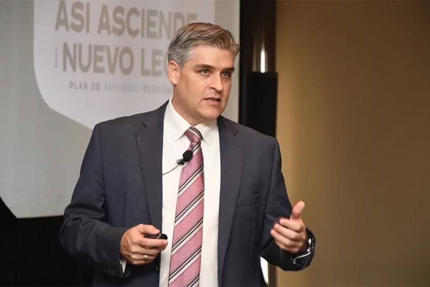 Iván Rivas con traje y corbata sosteniendo un control remoto de PowerPoint detrás de una pantalla de proyección con una diapositiva en español que dice: "De esta manera se levanta Nuevo León."