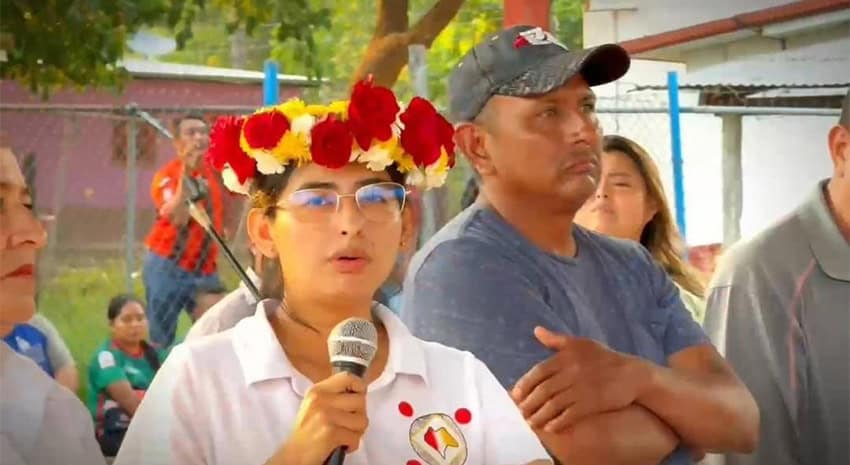 A young woman wearing glasses and a flower garland speaks into a microphone.