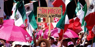 Anti-AMLO protesters wearing pink and waving Mexican flags crowd together with a sign saying "sin miedo, todos a votar," and bearing Xóchitl Gálvez's logo.