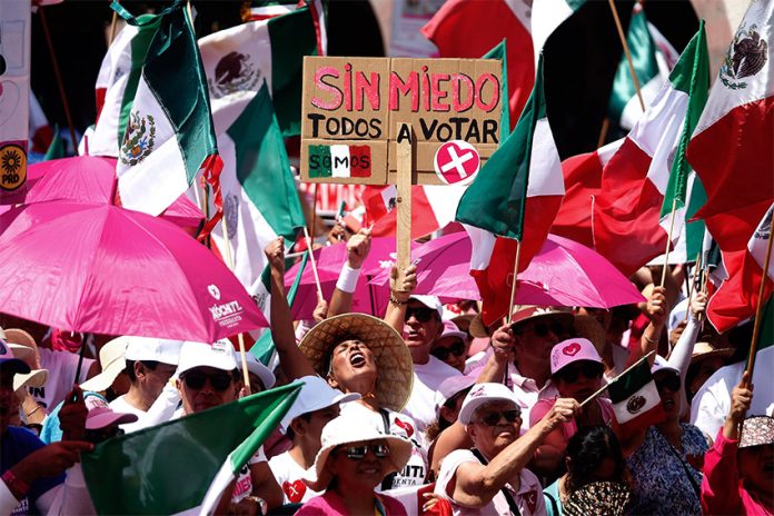 Anti-AMLO protesters wearing pink and waving Mexican flags crowd together with a sign saying 