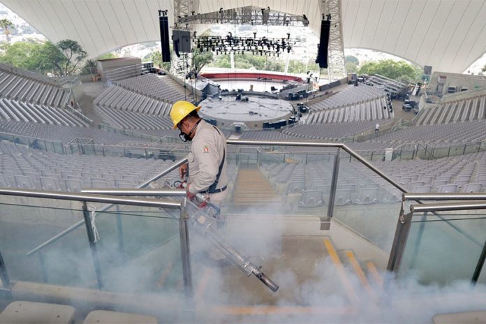 Man in uniform and hard hat spraying auditorium seats for mosquitos, surrounded by pesticide fumes.