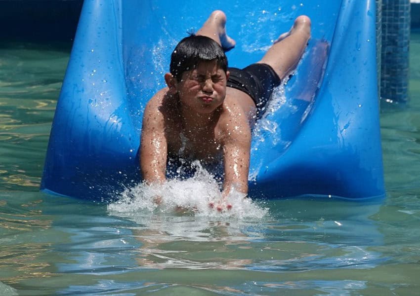 A Mexican boy in swim trunks landing head first in a swimming pool at the end of a blue plastic water slide