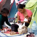 A migrant mother sitting in the entrance to a family sized green camping tent holds a nursing baby in one arm while she checks her cell phone. Her five-year-old daughter crouches in front of her. (Mireya Novo/Cuartoscuro)