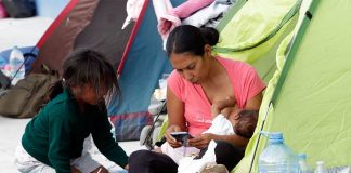 A migrant mother sitting in the entrance to a family sized green camping tent holds a nursing baby in one arm while she checks her cell phone. Her five-year-old daughter crouches in front of her. (Mireya Novo/Cuartoscuro)
