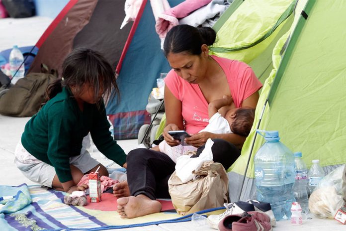 A migrant mother sitting in the entrance to a family sized green camping tent holds a nursing baby in one arm while she checks her cell phone. Her five-year-old daughter crouches in front of her. (Mireya Novo/Cuartoscuro)