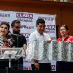 Mayor-Elect of Mexico City Clara Brugada at a podium holding an example of a construction brick made of recycled material. Four people next to her hold an example of recycled roofing material