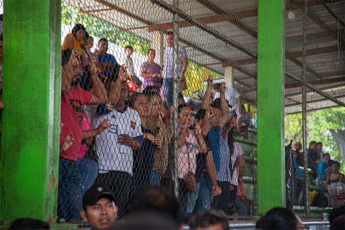 Refugees displaced by an armed attack on their Chiapas town stand in the bleachers of a open air sports court and look at proceedings below through a protective wire fence