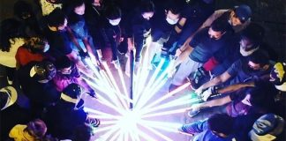 Members of the Jedi Knight Academy Mexico City at night gathered in a circle with each person extending their incandescent lightsabers to touch all together at the circle's center.