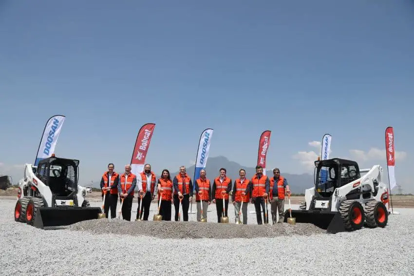 Government and company officials hold shovels and pose with Bobcat construction equipment at manufacturing plant groundbreaking ceremony.