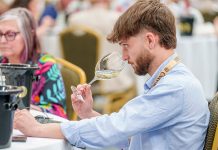 Concours Mondial de Bruxelles wine taster seated in a large hall in Leon, Mexico, samples a white wine from a wine glass.