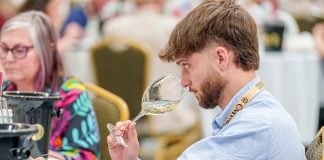 Concours Mondial de Bruxelles wine taster seated in a large hall in Leon, Mexico, samples a white wine from a wine glass.