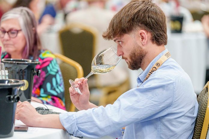 Concours Mondial de Bruxelles wine taster seated in a large hall in Leon, Mexico, samples a white wine from a wine glass.