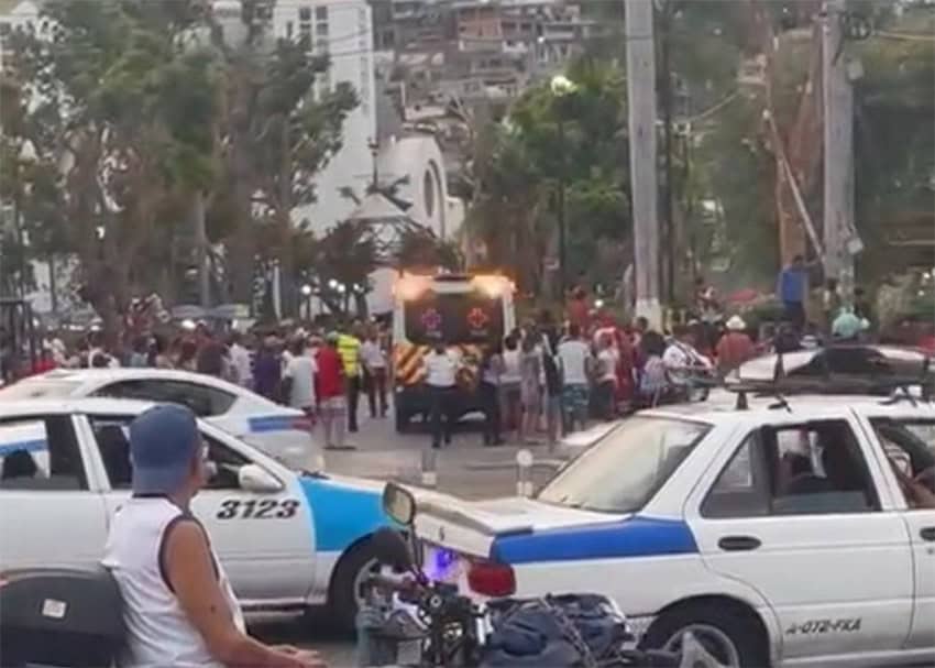 Emergency personnel load an injured person into an ambulance in a crowded public square in Acapulco with a cathedral in the background.