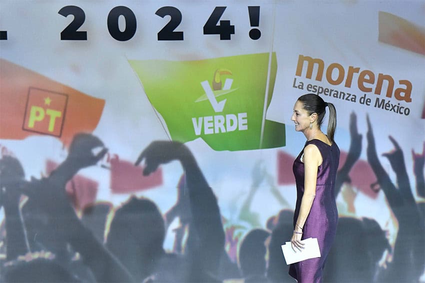 Claudia Sheinbaum walks across a stage with the logos of Morena, the PT and the PVEM parties behind her.