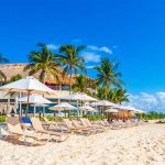 Beach loungers with parasols lying empty on a Playa del Carmen beach with palm trees and small amounts of seaweed in the background on the shore.