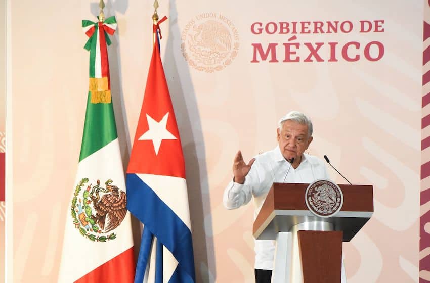 President Lopez Obrador at a podium gesturing with his right hand open, standing next to a flag of Mexico and a Cuban flag side by side.